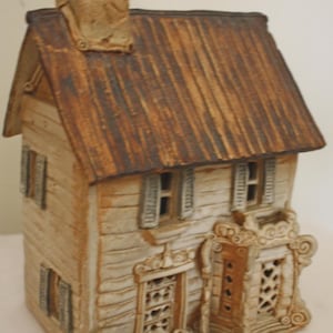 May include: A hand-crafted ceramic house with a brown roof, white walls, and detailed windows and doors. The house has a whimsical, cottage-like style.