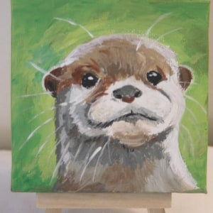 May include: A square canvas painting of an otter head featuring brown, white, and gray fur tones against a bright green background. The otter has dark eyes, a small nose, and long white whiskers painted with visible brushstrokes on a wooden easel.