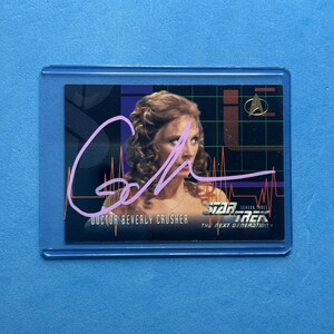 May include: A trading card featuring Doctor Beverly Crusher from Star Trek: The Next Generation. The card has a photo of the character, a signature, and the Star Trek logo. The card is encased in a protective plastic sleeve.