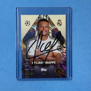 May include: A Topps trading card featuring Kylian Mbappé in a white Real Madrid jersey, encased in a clear plastic sleeve. The card displays a black ink autograph across the centre, the player's name, team logos, and statistical ratings of 33 and 97.
