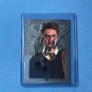 Op de afbeelding: Een rechthoekige verzamelkaart met een portret van een persoon met bruin haar, gekleed in een donkere trui over een wit overhemd met kraag en een das met patroon. De kaart heeft een metallic zilveren rand en een handtekening in zwarte inkt.