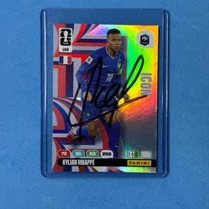 Puede incluir: Una tarjeta coleccionable Panini FIFA que muestra a Kylian Mbappé con la equipación azul de la selección francesa. La tarjeta tiene un acabado holográfico, un autógrafo en tinta negra y muestra el texto Kylian Mbappé, Icon, 180, con estadísticas 72, 91, 93, 256.