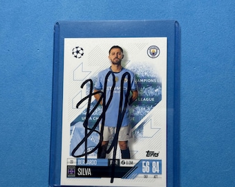 May include: A Bernardo Silva Topps trading card, featuring a player in a light blue jersey. The card is encased in a protective sleeve. The card has the text "Champions League" and "Topps". The card is signed.