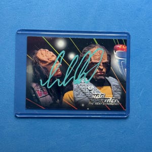 May include: A Star Trek: The Next Generation trading card featuring two characters with alien features. The card is encased in a clear plastic sleeve. The card has an autograph in light blue ink.