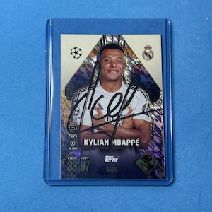 Puede incluir: Una tarjeta coleccionable Topps de Kylian Mbappé con la camiseta blanca del Real Madrid, dentro de una funda de plástico transparente. La tarjeta muestra una firma negra, el logotipo de la Champions League y estadísticas de 33 DEF y 97 ATT.