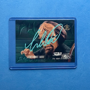 May include: A Star Trek: The Next Generation trading card featuring Lieutenant Worf. The card has a teal signature and is encased in a protective sleeve. The card's background is green with the Star Trek logo.