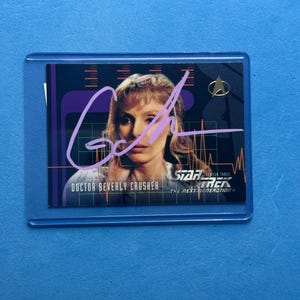 May include: A Star Trek: The Next Generation trading card featuring Doctor Beverly Crusher. The card has a purple signature across the image. The card is in a protective sleeve. The background is a solid blue.