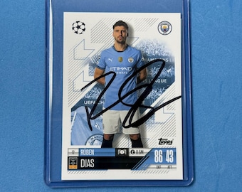 May include: A trading card of soccer player Rúben Dias in a protective sleeve. The card features a photo of Dias in a light blue jersey with the Etihad Airways logo. The card is signed and includes the Topps logo and player statistics.