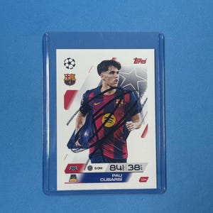 Puede incluir: Una tarjeta coleccionable Topps de Pau Cubarsí en una funda protectora azul. La tarjeta muestra al futbolista con una camiseta azul oscuro y roja. Incluye el logotipo de Topps, los logotipos del equipo y estadísticas del jugador.