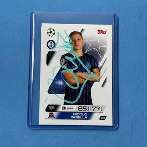 Pode incluir: Um cartão colecionável Topps com Nicolo Barella com uma camisa azul e preta. O cartão está dentro de uma manga protetora de plástico. O logotipo da Topps e as estatísticas do jogador são visíveis.