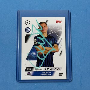 Pode incluir: Um cartão de negociação de futebol de Nicolo Barella em uma manga protetora. O cartão apresenta o jogador em uma camisa preta e azul, com uma assinatura turquesa. O cartão tem o logotipo da Topps e estatísticas do jogador.