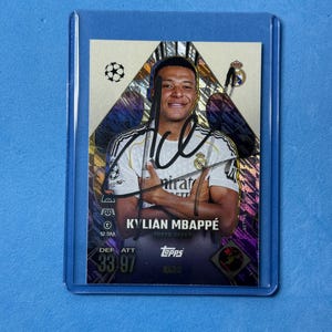 Puede incluir: Una tarjeta coleccionable Topps con una fotografía de Kylian Mbappé vistiendo una camiseta blanca del Real Madrid con los brazos cruzados. La tarjeta tiene un fondo holográfico morado y dorado, un autógrafo en tinta negra y texto que incluye Kylian Mbappé, Super Speed y 33/97.