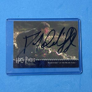 Puede incluir: Una tarjeta coleccionable de Harry Potter en una funda protectora, con una escena y el texto "Dementors at the Black Lake". La tarjeta tiene una firma con tinta negra.