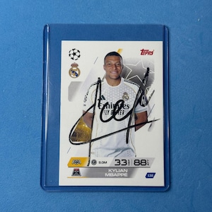 Può includere: Una figurina Topps con una fotografia di Kylian Mbappé che indossa una maglia bianca del Real Madrid con il logo Emirates Fly Better. La carta include un autografo a inchiostro nero, lo stemma del Real Madrid e alcune statistiche.