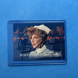 May include: A Star Trek: The Next Generation trading card featuring Doctor Beverly Crusher in a nurse's uniform. The card has an autograph and the Star Trek logo. The background is blue.