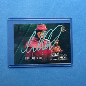 May include: A Star Trek: The Next Generation trading card featuring Lieutenant Worf in a red uniform and hat. The card is signed in blue ink and is encased in a protective sleeve. The card also has the Star Trek logo.