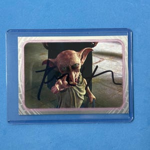 Puede incluir: Una tarjeta coleccionable que representa a un personaje de una película de fantasía. La tarjeta muestra una criatura de piel clara con orejas grandes y una nariz larga, vestida con una prenda de color beige. La tarjeta está dentro de una funda protectora de plástico, con un fondo azul.