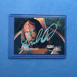 May include: A Star Trek: The Next Generation trading card featuring Lieutenant Worf. The card has a teal signature across the image. The card is in a protective sleeve against a blue background.