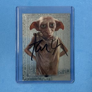 Puede incluir: Una tarjeta coleccionable con un personaje de orejas grandes y ojos verdes, firmada con tinta negra. La tarjeta está dentro de una funda protectora de plástico, sobre un fondo azul claro. El personaje es de la serie Harry Potter.