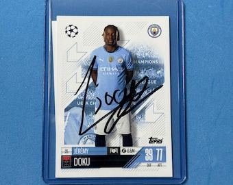 May include: A Topps trading card of soccer player Jeremy Doku in a protective sleeve. The card features Doku in a light blue jersey with the Etihad Airways logo. The card is signed and includes the player's name, team logo, and statistics.