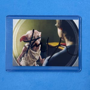 Pode incluir: Um cartão colecionável com um personagem de um filme de fantasia. O cartão mostra uma criatura com orelhas grandes e uma pessoa ao fundo. O cartão está em uma manga de plástico transparente, em um fundo azul.