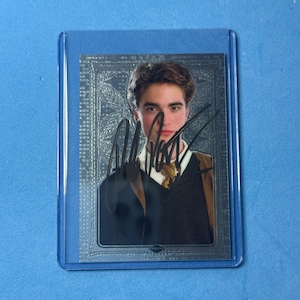 Puede incluir: Una tarjeta coleccionable rectangular dentro de una funda protectora de plástico transparente que muestra el retrato de una persona de cabello castaño con suéter oscuro, camisa blanca y corbata estampada, con una firma en tinta negra.