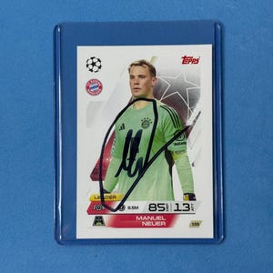 Può includere: Una figurina Topps con una fotografia di Manuel Neuer che indossa una maglia da portiere verde chiaro. La carta include il logo dell'FC Bayern Monaco, l'emblema della Champions League e un autografo a inchiostro nero al centro. È racchiusa in una custodia protettiva in plastica trasparente.