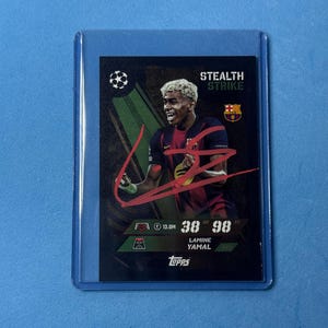 Puede incluir: Una tarjeta coleccionable Topps que muestra a Lamine Yamal con la camiseta del FC Barcelona y un autógrafo rojo en el centro. La tarjeta negra presenta el texto Stealth Strike, el logo de la Champions League y estadísticas de 38 DEF y 98 ATT.