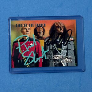 May include: A Star Trek: The Next Generation trading card in a protective sleeve. The card features three characters from the episode "Sins of the Father" and includes autographs. The card is set against a blue background.