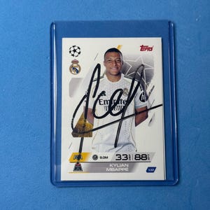 Puede incluir: Una tarjeta coleccionable de Topps que muestra a Kylian Mbappé con una camiseta blanca del Real Madrid y un autógrafo en tinta negra en el frente. La tarjeta exhibe el logo de la Champions League, el escudo del Real Madrid y estadísticas de jugador.