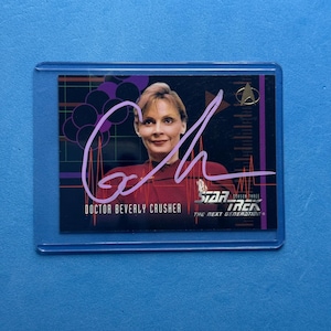 May include: A Star Trek: The Next Generation trading card featuring Doctor Beverly Crusher. The card has a purple signature and the Star Trek logo. The background is black with purple and white accents. The card is in a protective sleeve.