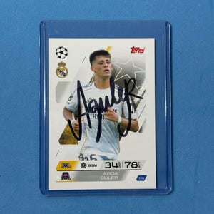May include: A trading card featuring a soccer player in a white jersey with a black signature. The card has the Topps logo and the Real Madrid logo. The card is in a protective sleeve.
