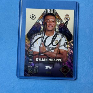 May include: A Topps trading card featuring Kylian Mbappé in a white Real Madrid jersey with a black signature across the front. The card has a gold and purple geometric background, the Champions League logo, and player stats including 33 DEF and 97 ATT.