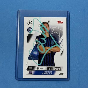 Pode incluir: Um cartão colecionável de futebol Topps de Nicolo Barella em uma manga protetora. O cartão apresenta uma foto do jogador em uma camisa preta e azul. O cartão tem o logotipo da Topps e as estatísticas do jogador. O cartão está em uma manga de plástico transparente.