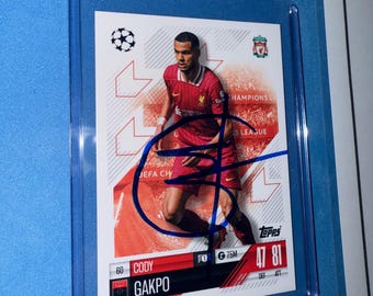 Może przedstawiać: Karta kolekcjonerska Topps z Codym Gakpo, piłkarzem Liverpoolu FC. Karta jest czerwona, biała i niebieska ze zdjęciem Gakpo w czerwonej koszulce. Karta ma niebieski podpis i tekst "Cody Gakpo" i "47 81".