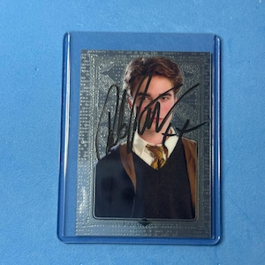 Op de afbeelding: Een rechthoekige verzamelkaart met een portret van een persoon met bruin haar, gekleed in een wit overhemd met kraag, een geel-zwart gestreepte das en een donker vest. De kaart heeft een zilverkleurige rand en een handtekening in zwarte inkt.