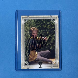 Könnte beinhalten: Sammelkarte mit einer Person in gelber und marineblauer Uniform, mit einem Autogramm in schwarzer Tinte. Die Karte befindet sich in einer transparenten Kunststoffhülle auf blauem Hintergrund.