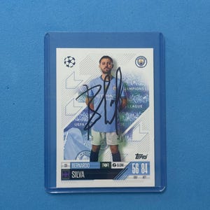 Pode incluir: Um cartão colecionável de Bernardo Silva em uma manga protetora. O cartão apresenta o jogador de futebol em uma camisa azul clara. O texto inclui "Champions League", "UEFA", "Bernardo Silva" e "Topps". O cartão é assinado.