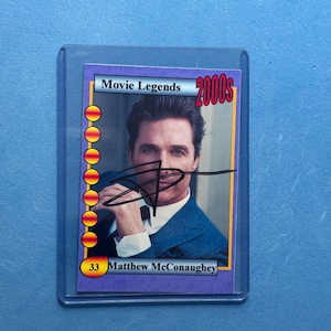 May include: A collectible trading card featuring Matthew McConaughey from the Movie Legends 2000s series. The card has a purple border with gold accents and the actor's signature. The card is encased in a protective plastic sleeve.