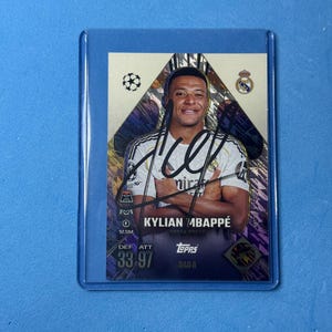May include: A Topps trading card featuring Kylian Mbappé in a white Real Madrid jersey with a black ink autograph across the centre. The card has a metallic gold and purple geometric background, the Champions League logo, and stats showing 33 DEF and 97 ATT.