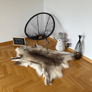 Op de afbeelding: Een rendierhuidkleed met een mix van bruin, grijs en wit bont wordt op een houten vloer getoond. Een zwarte stoel met een cirkelvormige geweven rug en metalen poten staat op de achtergrond. Een klein schoolbord met het nummer 253 is ook zichtbaar.