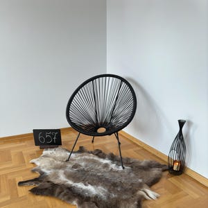 Könnte beinhalten: Ein schwarzer Stuhl mit geflochtenem Design steht auf einem hellbraunen Holzboden mit einem weißen Kunstfellteppich. Eine schwarze Digitaluhr mit der Zeit 6:57 steht auf dem Boden neben dem Stuhl. Eine schwarze Metallvase mit einer Kerze darin steht in der Ecke des Raumes.