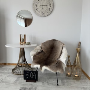 Op de afbeelding: Een witte stoel met een bruin en wit schapenvacht erop. De stoel staat in een kamer met een witte muur, een ronde spiegel, een witte tafel met een gouden metalen onderstel en twee gouden metalen kaarsenstandaards. Een zwart schoolbord met het nummer 604 erop staat op de voorgrond.