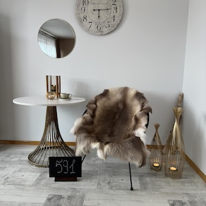 Op de afbeelding: Een kamer met een ronde witte tafel met een gouden voet, een zwarte stoel met een vacht en decoratieve gouden kandelaars. Een grote wandklok en een kleine spiegel zijn ook zichtbaar.