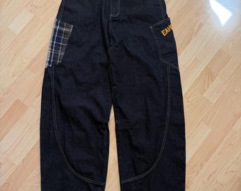 May include: Dark wash denim jeans with a relaxed fit. Features a plaid patch on the left leg and a pocket with the letters "EAH" in gold. The jeans have light stitching and a button closure.