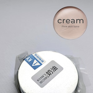 May include: A cream-coloured jar with a white lid, sealed in clear plastic. The label reads "KYAN'S WORKS 奶油". Above, a circular swatch displays "cream" and "Pink skin tone".