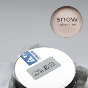May include: A round, silver-lidded container of light skin tone makeup labelled "snow". The product is packaged in clear plastic. The text "KYAN'S WORKS 超白" is printed on a label on the container.