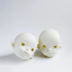 Può includere: Due teste di bambola bianche con orbite gialle su una superficie bianca. Le teste hanno una consistenza liscia e un'espressione leggermente malinconica. Le orecchie sono dettagliate e l'estetica generale è minimalista.