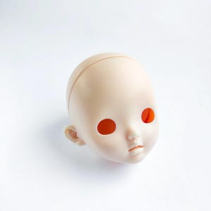 May include: A blank doll head in a light beige color, with large, orange eye sockets and a small, defined mouth. The head has a smooth surface and visible ear details. The doll head is a blank canvas for customization.