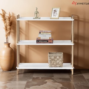May include: A three-tiered white shelf unit with light wood supports. The shelves hold a vase with dried plants, books, a woven basket, and decorative items. The background is a warm beige wall with arched alcoves.